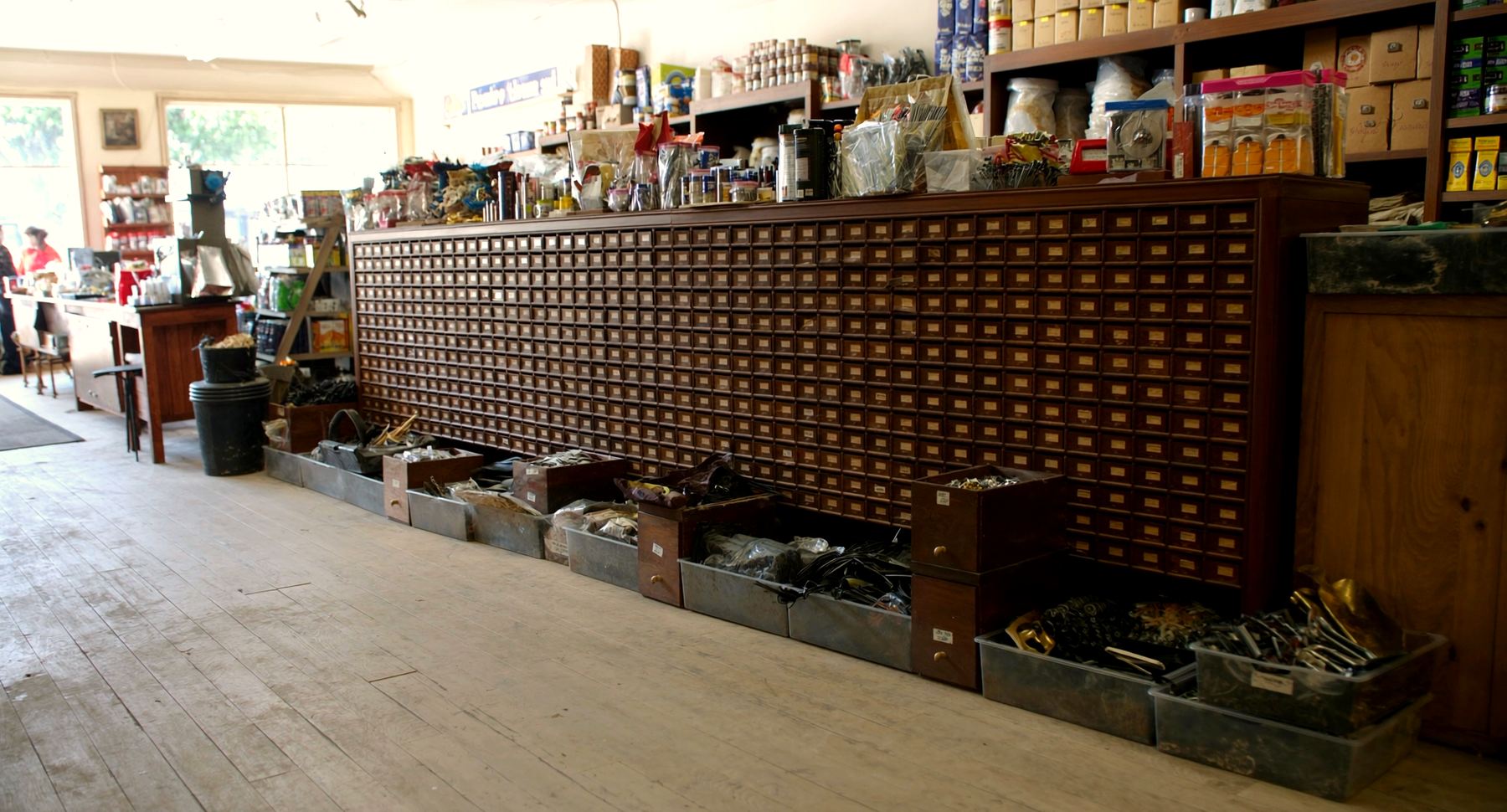 A wall of small wooden bin drawers with brass label holders running the length of the store, with open metal trays of loose hardware set along the floor in front of it.