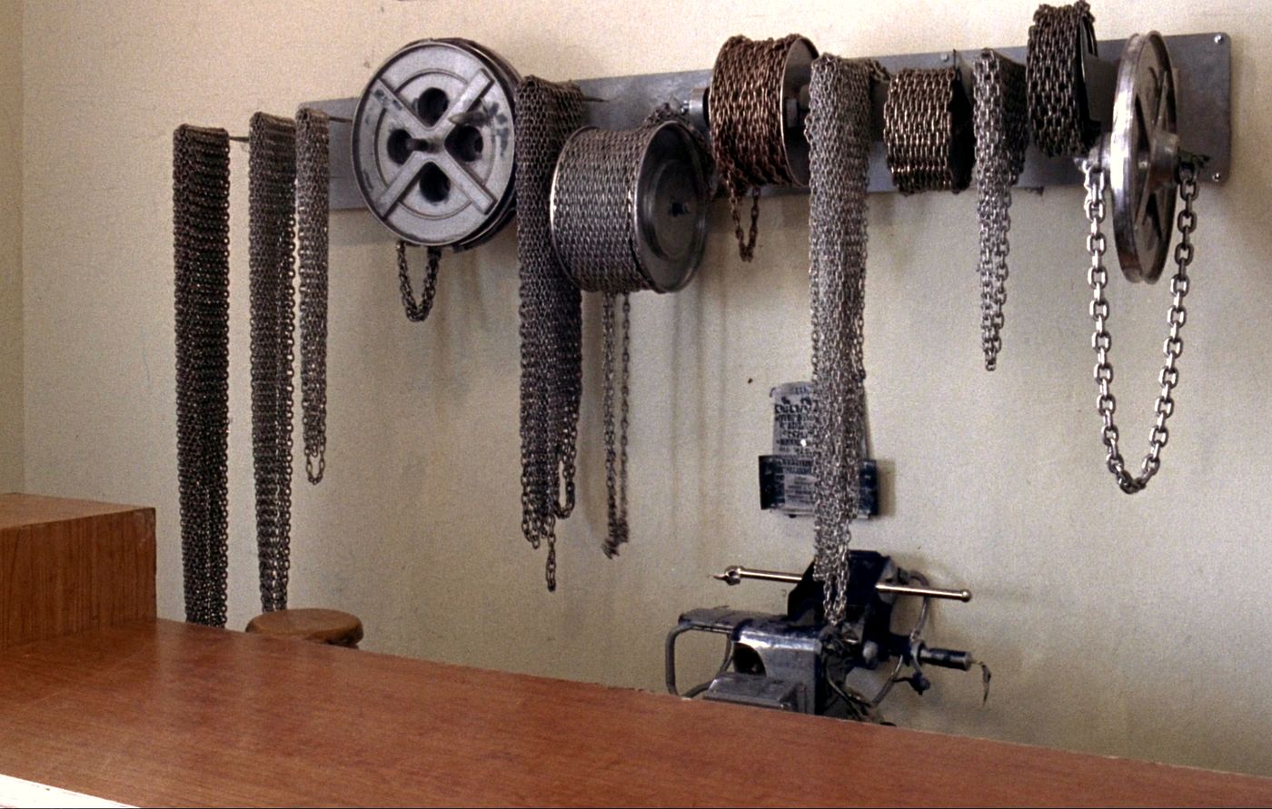 Eight chain reels bolted to a steel rail on the wall above the counter, each carrying a different link size hanging loose, with a bench vise mounted below for cutting.