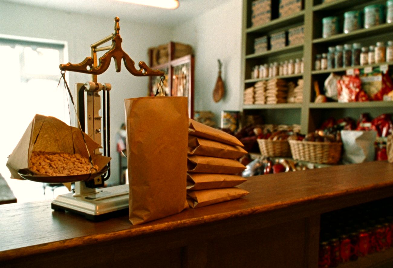 A brass balance scale on the wooden counter with an open paper sack of dry beans resting on the weighing pan, a row of filled and folded paper sacks stacked beside it, and stocked shelves behind.