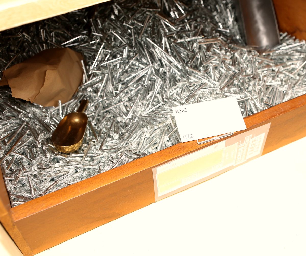 An open wooden bin of galvanized nails with a brass scoop lying in it, a brown paper sack tucked against one side, and a small white card in a wire holder clipped to the front rail.