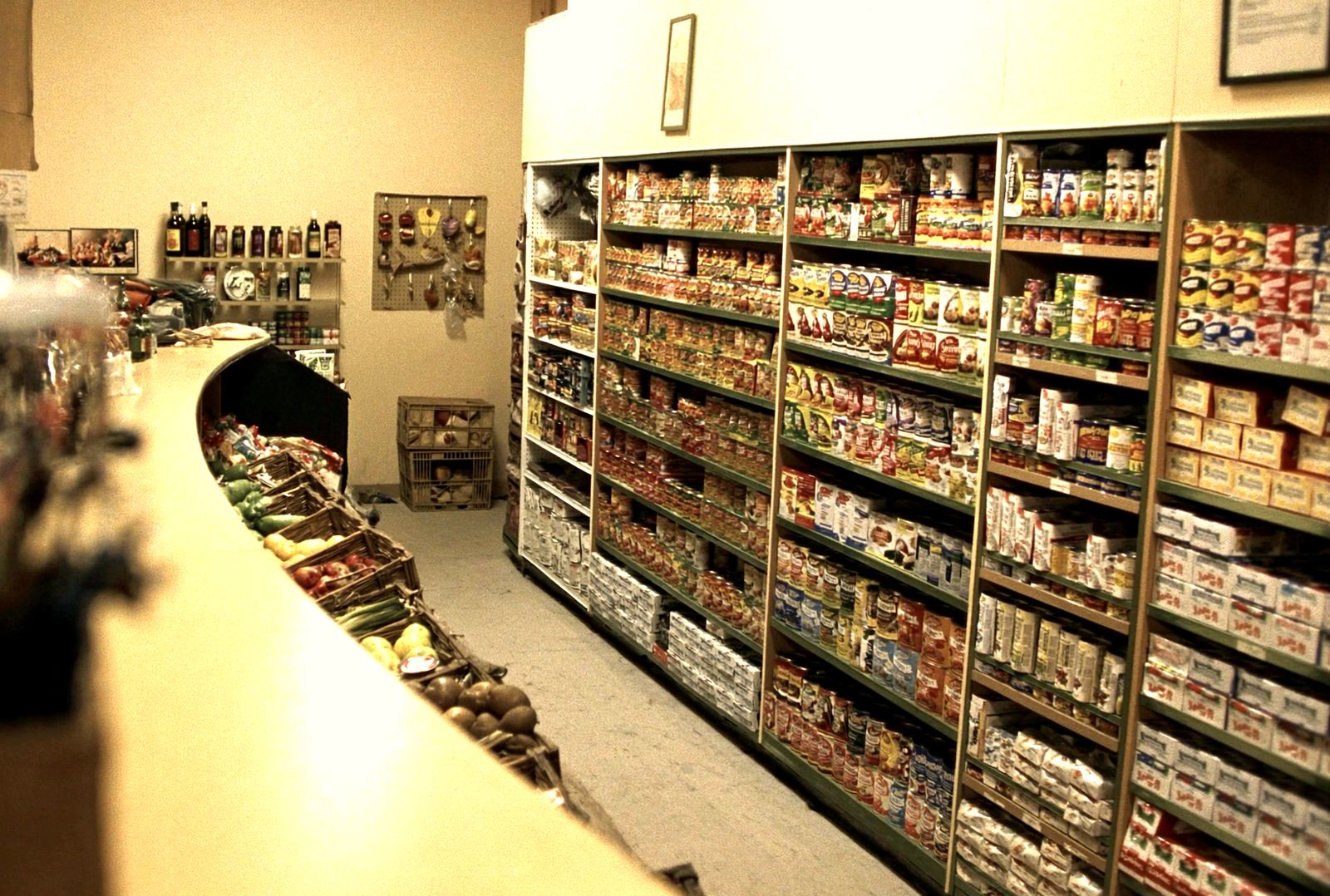Grocery shelves stocked deep with canned and boxed goods down the length of the aisle, with a curved wooden produce rack running along the other side.
